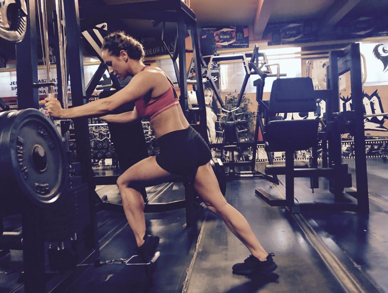 Kobieta w sportowym stroju ćwiczy na maszynie do nóg w siłowni, widoczne obciążenie i metalowa konstrukcja urządzenia.