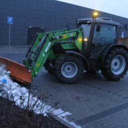 Zielony traktor z pomarańczowym pługiem odśnieżającym, zgarniający śnieg na tle szarego budynku z niebieskim znakiem parkingu.