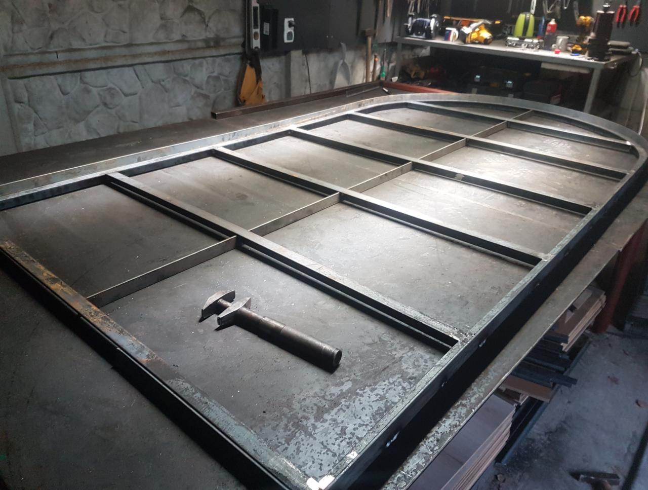 Metalowa konstrukcja okienna z łukiem na warsztatowym stole, obok młotek. Konstrukcja spawana, surowa, przygotowana do dalszej obróbki.