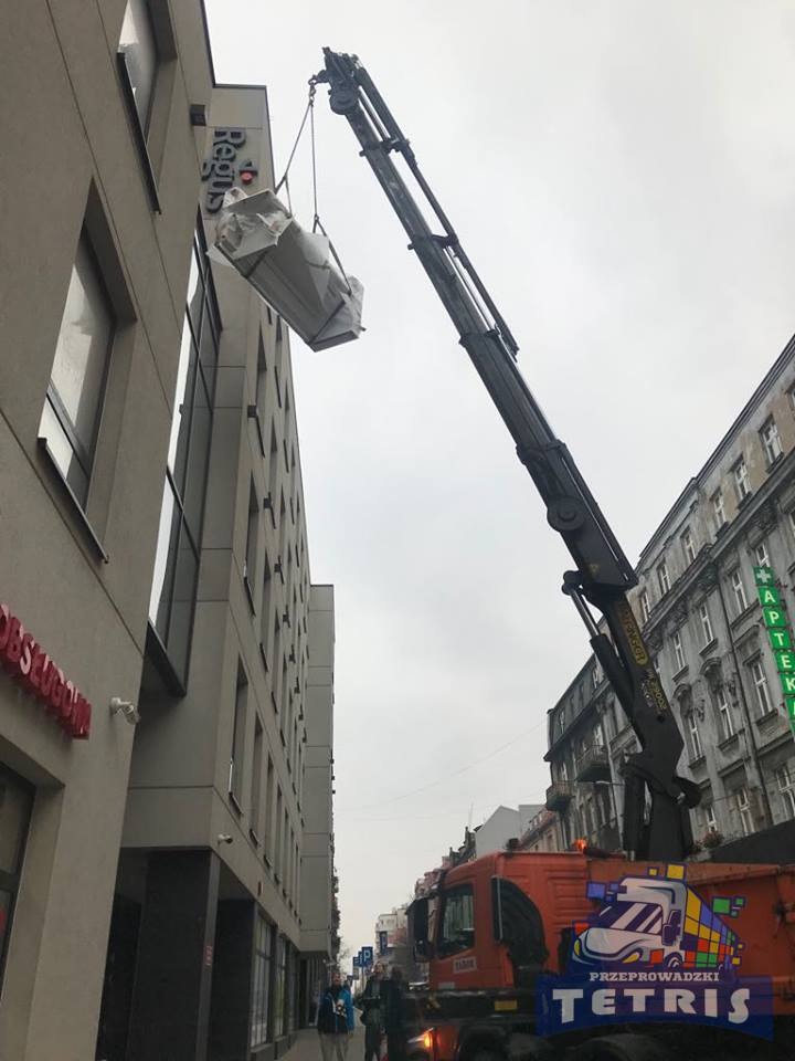 Żuraw samochodowy podnosi zabezpieczony ładunek na tle miejskiej zabudowy, widoczny budynek z napisem Regus, na pojeździe logo firmy Tetris.