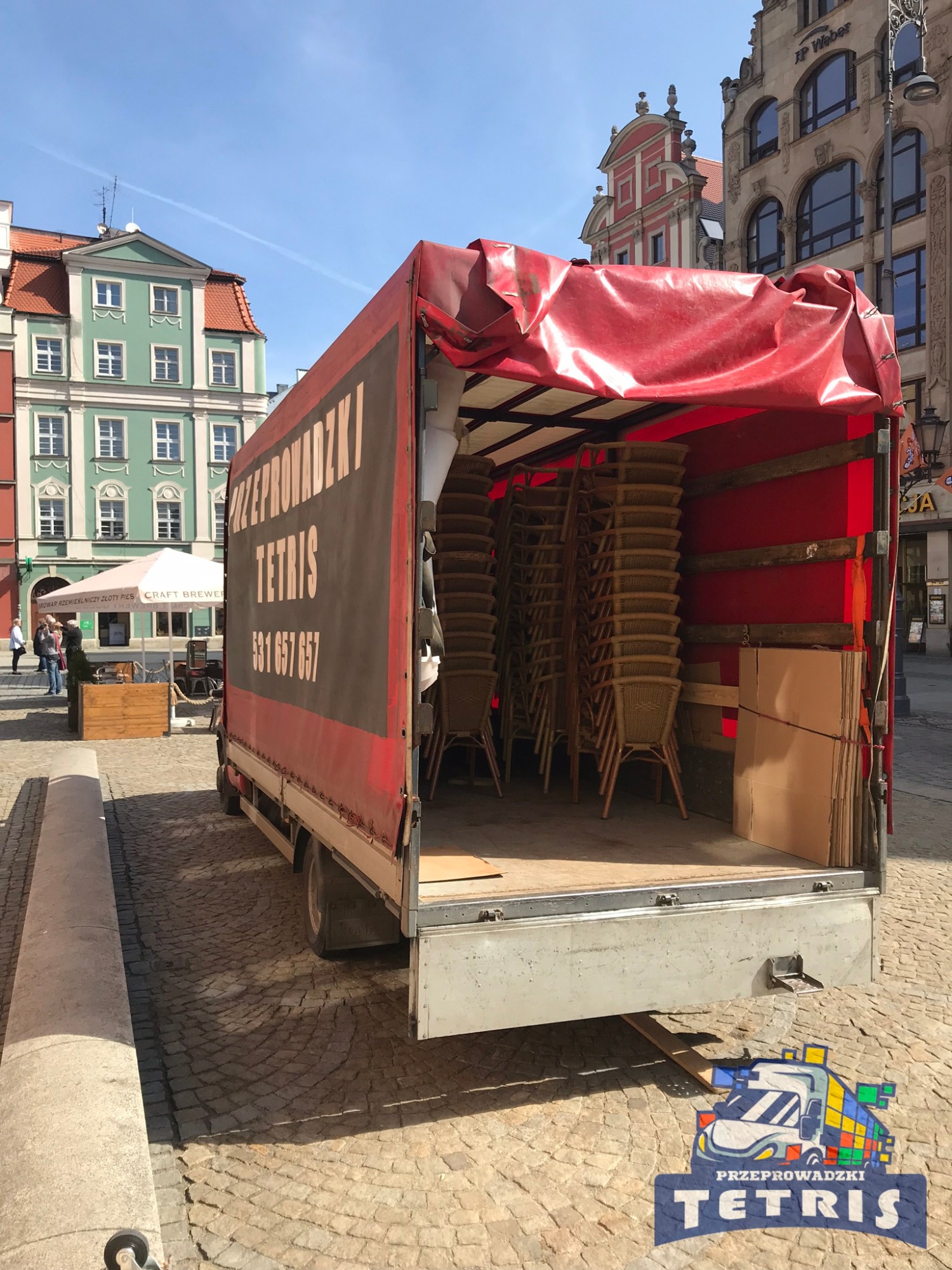 Otwarta paka samochodu dostawczego firmy przeprowadzkowej Tetris wypełniona stosem wiklinowych krzeseł i kartonami na tle brukowanej ulicy w historycznej części miasta.