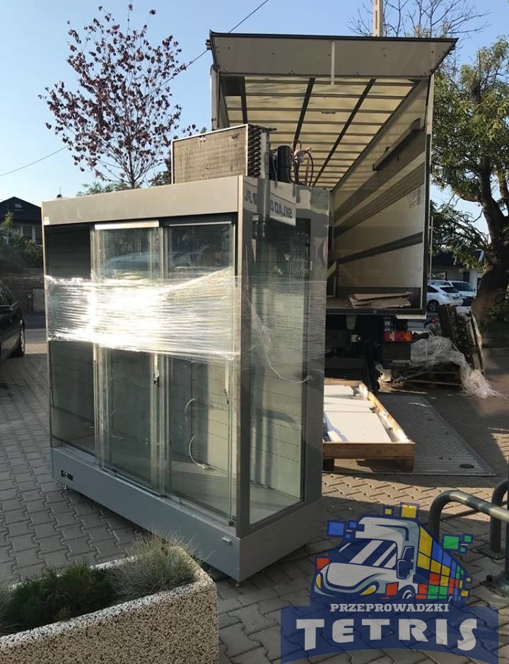 Srebrna chłodnicza witryna owinięta folią stretch, przygotowana do załadunku na samochód dostawczy z otwartą paką, widok z boku na chodniku.