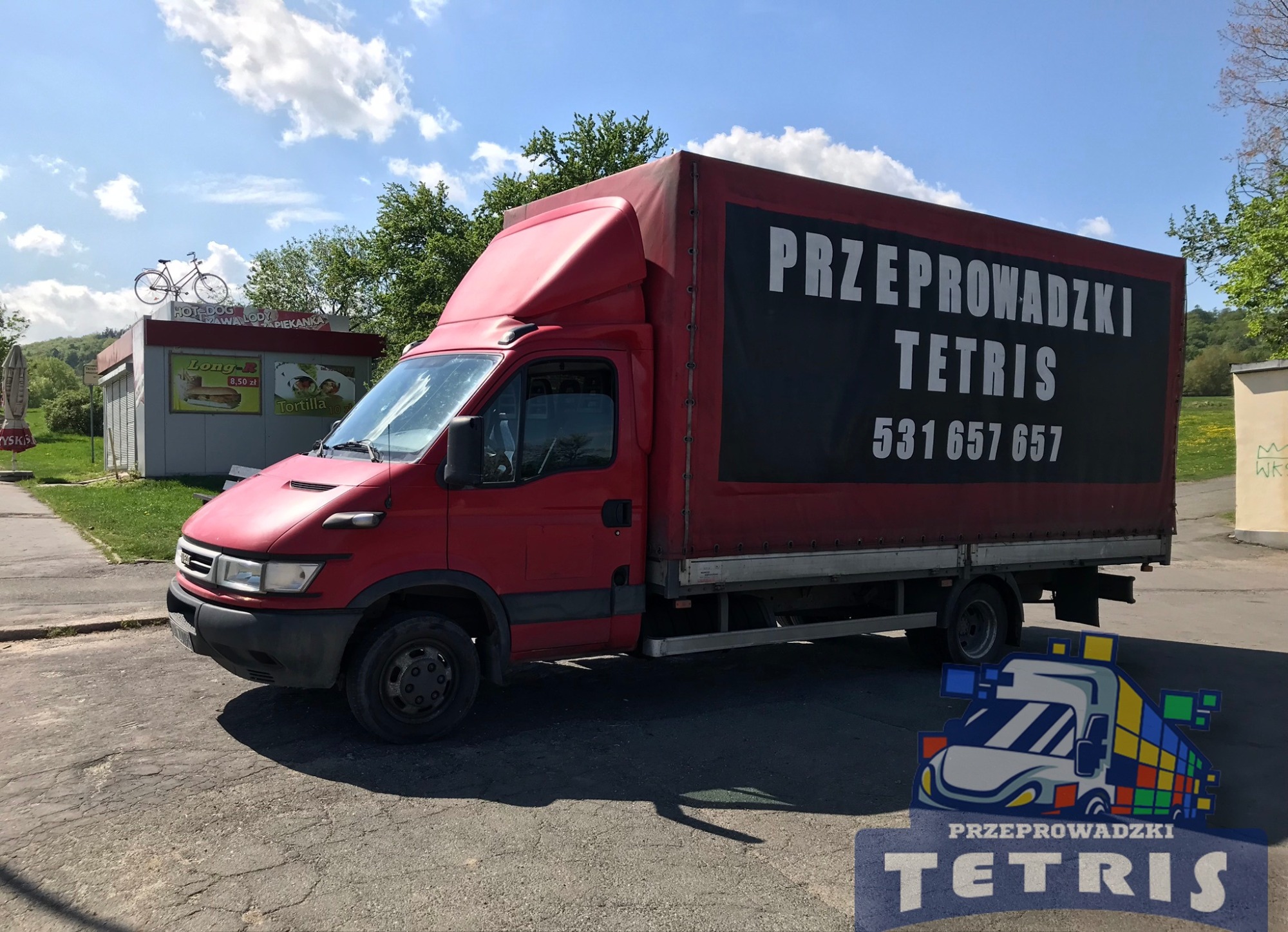 Czerwony samochód dostawczy z napisem 'Przeprowadzki Tetris' i numerem telefonu na plandece, zaparkowany na ulicy, z budką gastronomiczną i rowerem na dachu w tle. Na pierwszym planie logo firmy.