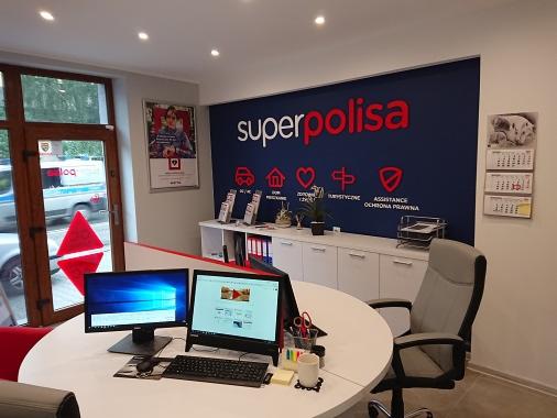 Wnętrze biura Superpolisa z dwoma monitorami na okrągłym stole, krzesłem biurowym i ścianą z logo firmy oraz symbolami: dom, samochód, serce, parasol, tarcza. Na ścianie kalendarz z kotami.