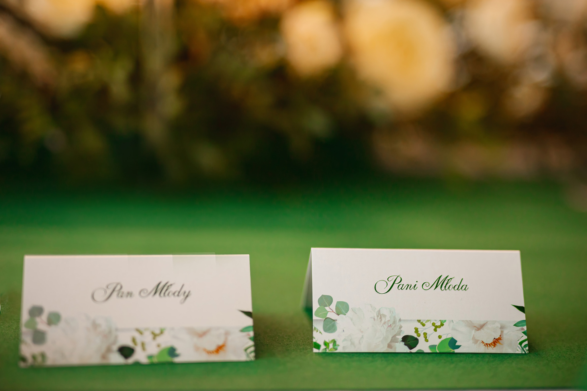Dwa wizytowniki ślubne z napisem 'Pan Młody' i 'Pani Młoda' z delikatnym kwiatowym wzorem na zielonym tle, ustawione na stole podczas uroczystości.