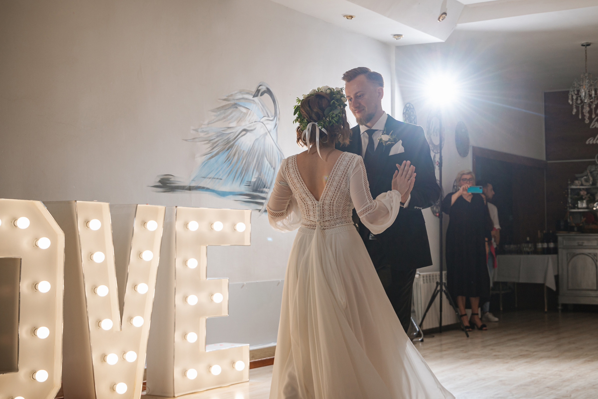Para młoda tańczy pierwszy taniec w sali z dekoracją LOVE z żarówkami, panna młoda w sukni z koronkowymi plecami i wiankiem, pan młody w czarnym garniturze, w tle malowidło łabędzia.