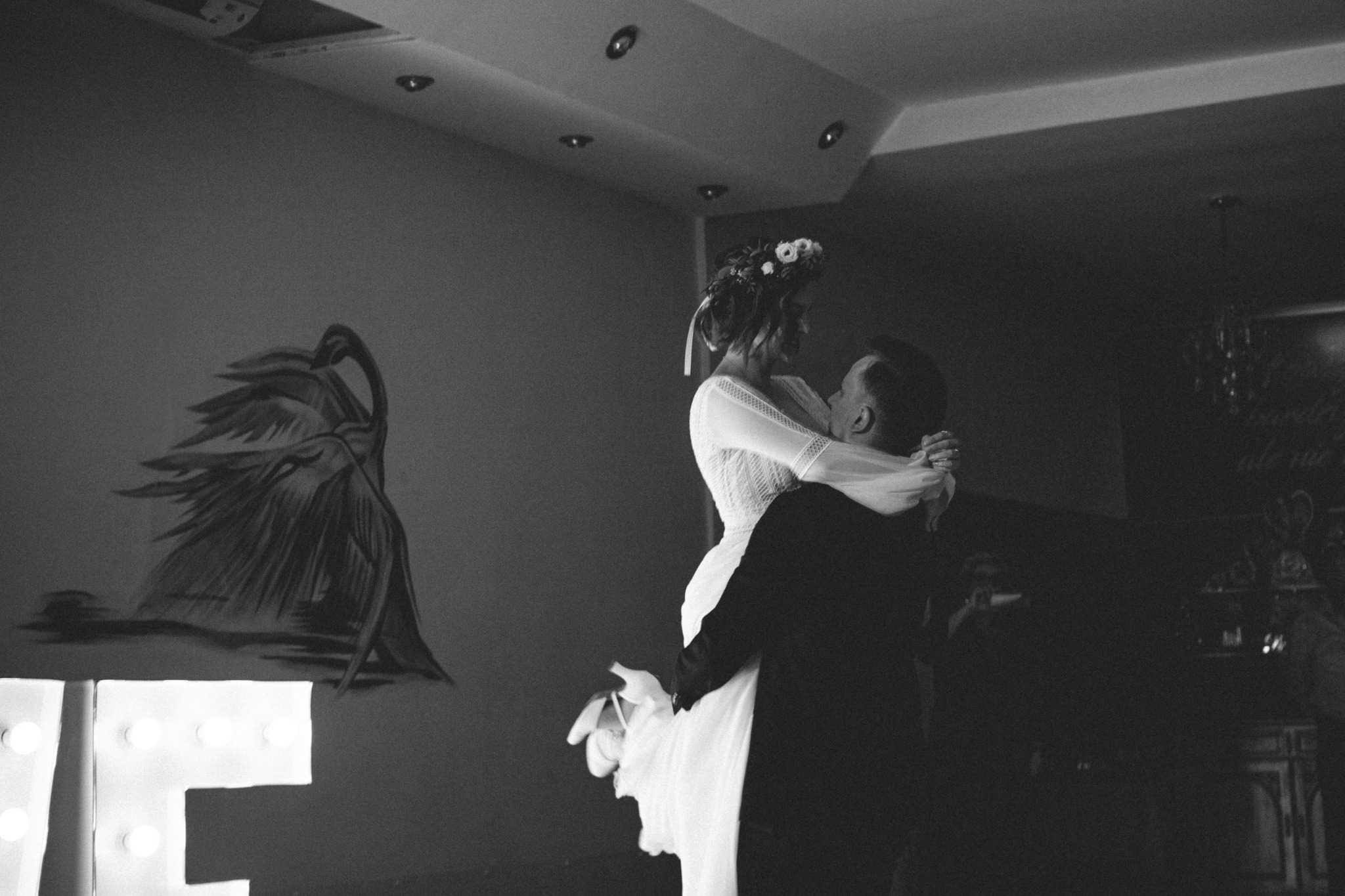 Czarno-białe zdjęcie nowożeńców podczas pierwszego tańca, panna młoda w koronkowej sukni z wiankiem na głowie, unoszona przez pana młodego na tle ściany z malowidłem i podświetlanymi literami.