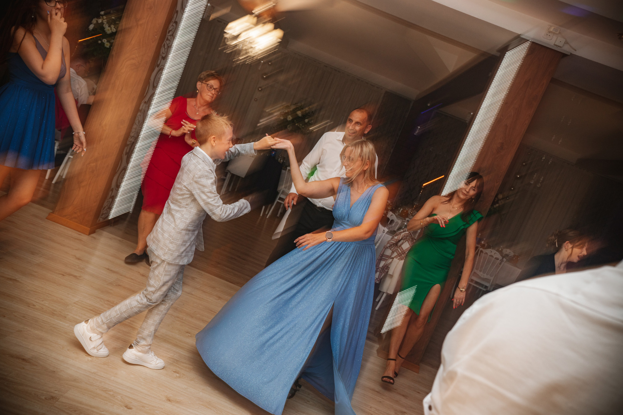 Rozmazane ujęcie tańczących gości w sali balowej, kobieta w długiej, błękitnej sukni z rozcięciem, chłopiec w szarej kraciastej marynarce i białych sneakersach, oświetlenie sufitowe w tle.