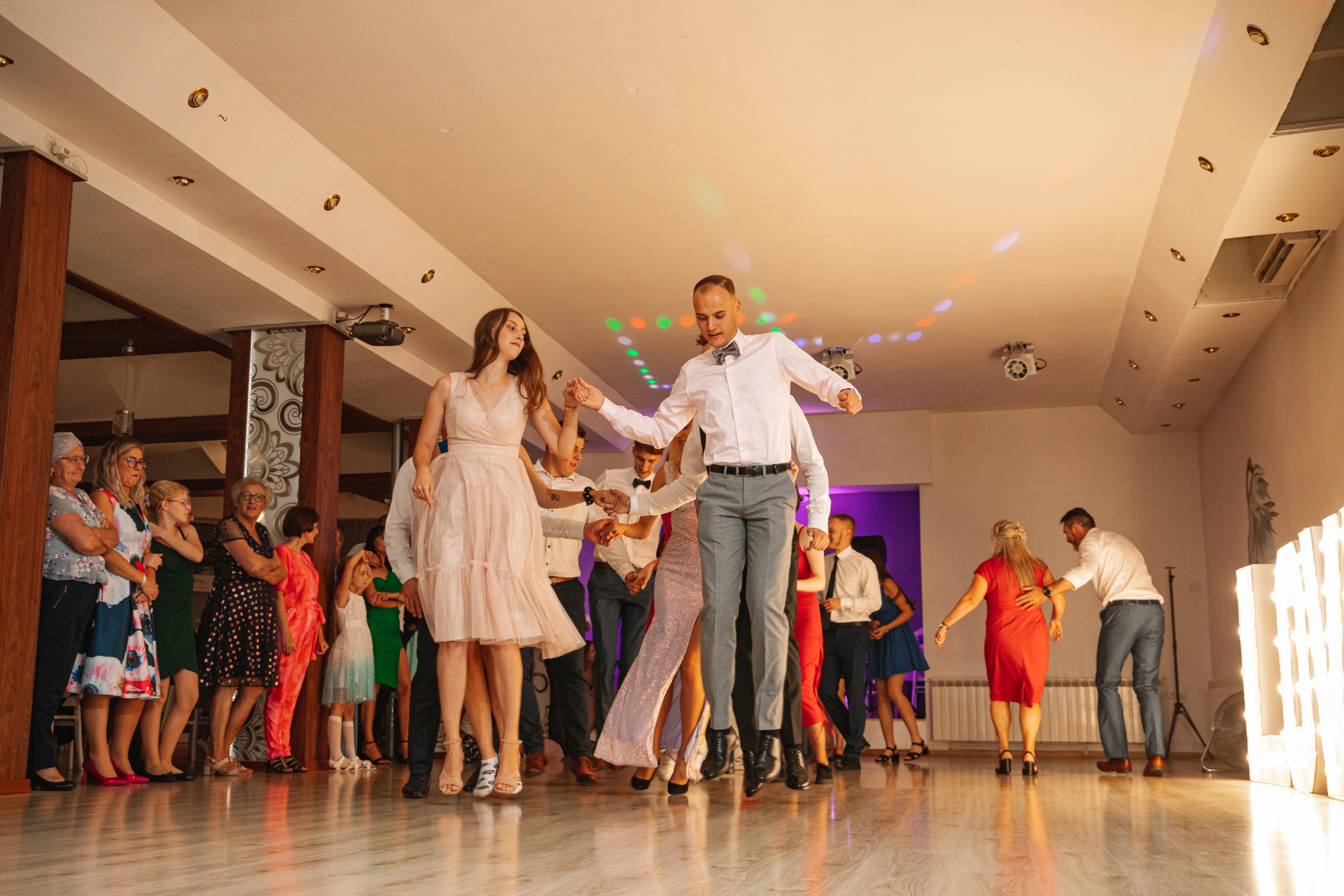 Grupa osób w różnym wieku, w strojach wizytowych, skacze i tańczy na parkiecie w sali balowej, oświetlonej kolorowymi reflektorami.