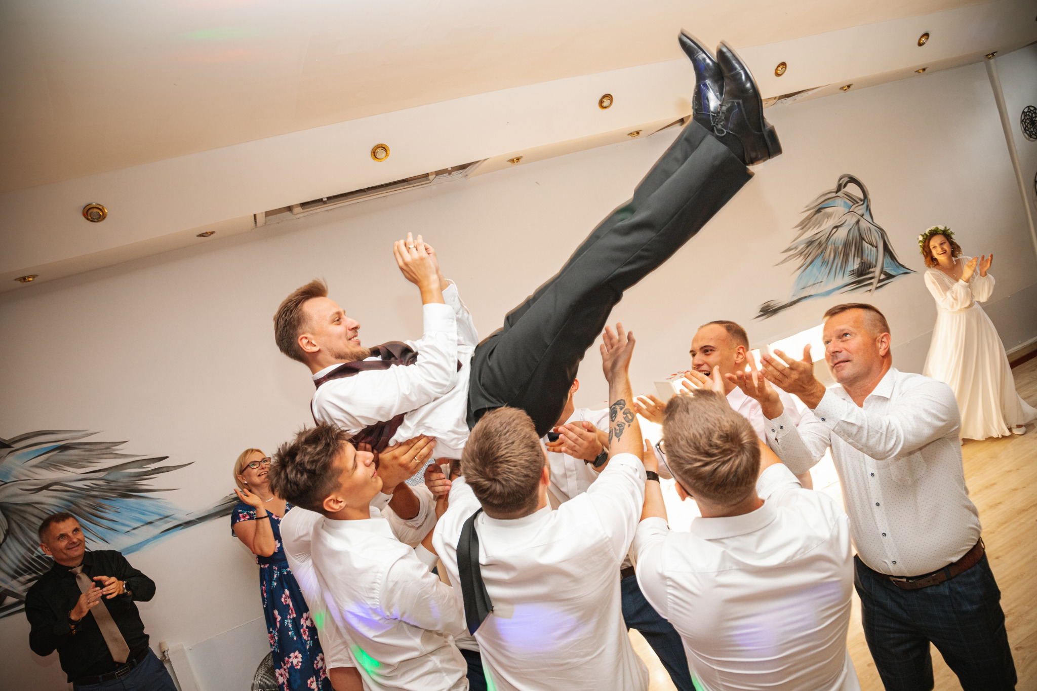 Groom being lifted by guests at a wedding reception, bride clapping in the background near a wall with a mural.
