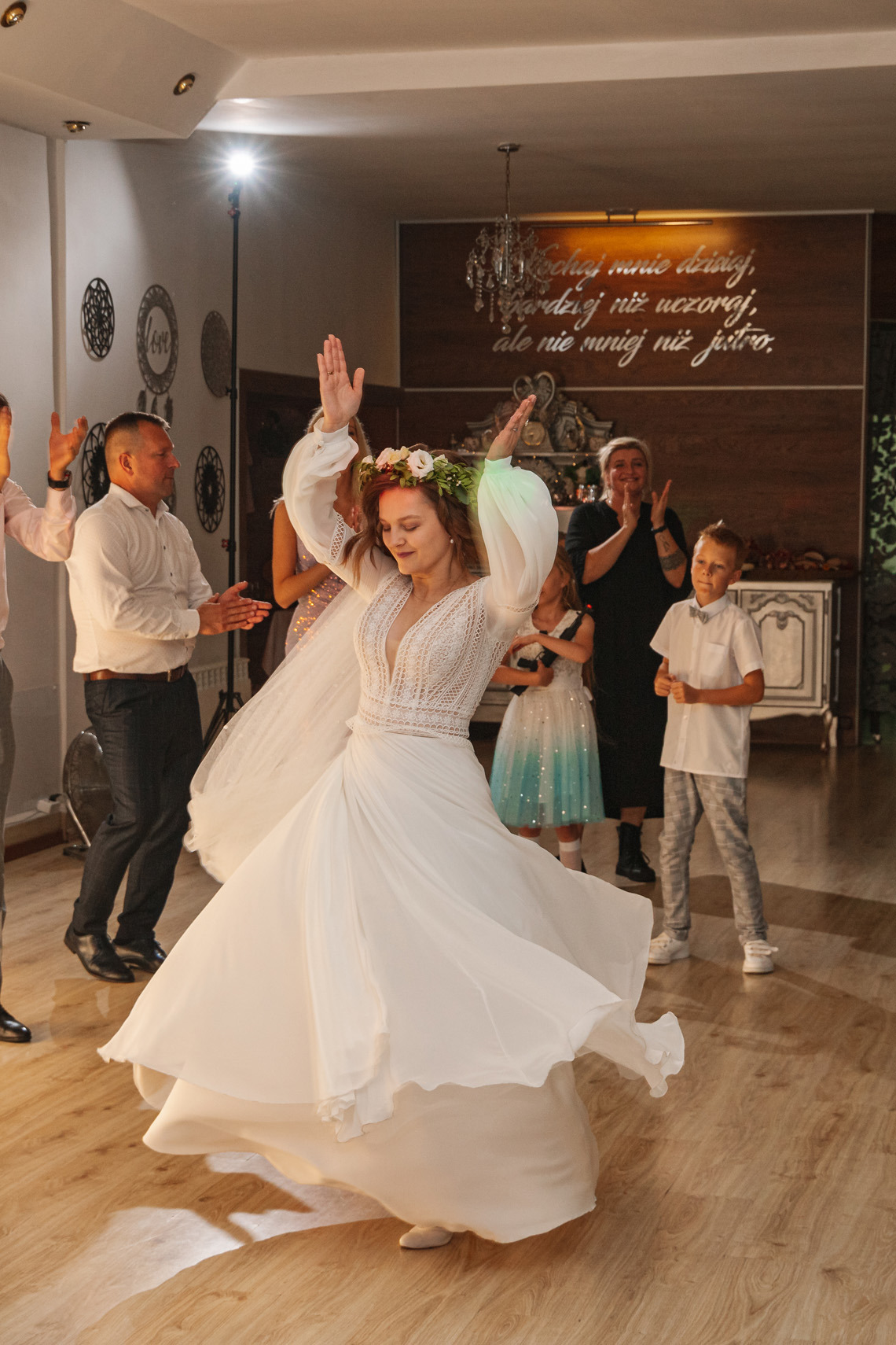 Panna młoda w białej sukni z wiankiem tańczy podczas wesela, w tle goście klaszczą i obserwują, oświetlenie sceniczne, drewniana podłoga.