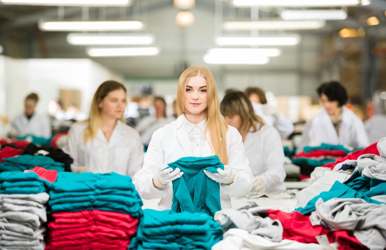 Pracownica w białym fartuchu i rękawiczkach sortuje zieloną koszulkę na tle stosów ubrań w hali produkcyjnej.