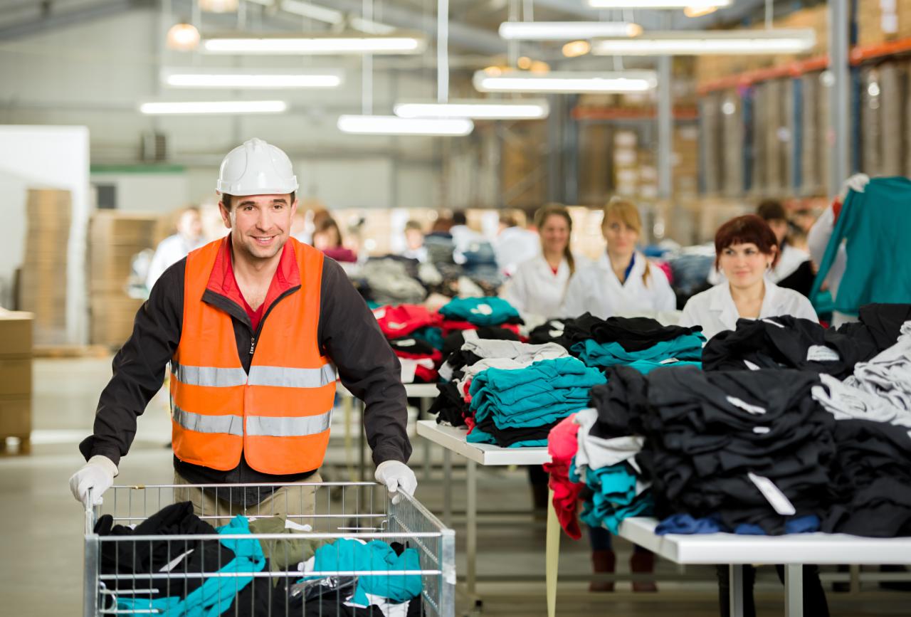 Pracownik w kasku i kamizelce odblaskowej pcha wózek z odzieżą w magazynie, w tle pracownicy przy stołach z ułożonymi ubraniami.