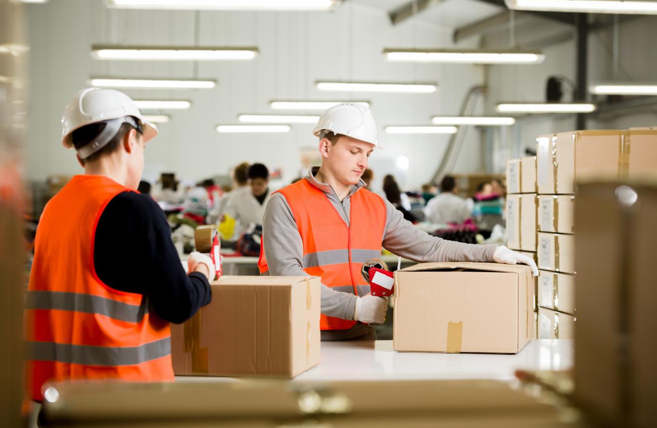 Pracownicy w magazynie, w kamizelkach odblaskowych i kaskach, zaklejają kartonowe pudełka taśmą za pomocą dyspensera, w tle widoczne inne osoby i przygotowane paczki.