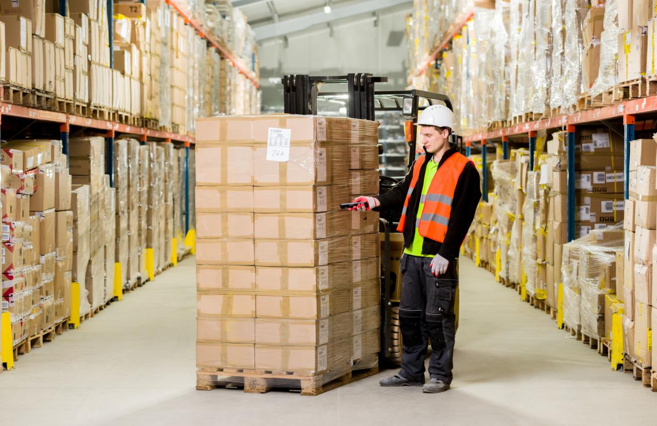 Pracownik magazynu w kasku i kamizelce odblaskowej skanuje kod kreskowy na palecie z zapakowanymi kartonami przy użyciu wózka widłowego w hali magazynowej.