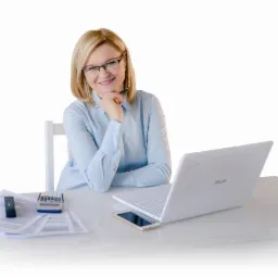 Uśmiechnięta blondynka w okularach siedzi przy biurku z laptopem, kalkulatorem, dokumentami i smartfonem, trzymając długopis przy brodzie w zamyślonej pozie.