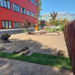 ARTI GARDEN - Rozwinięty fragment trawy z rolki na przygotowanym podłożu, obok leżą zwinięte rolki, w tle czerwony budynek i taczka.