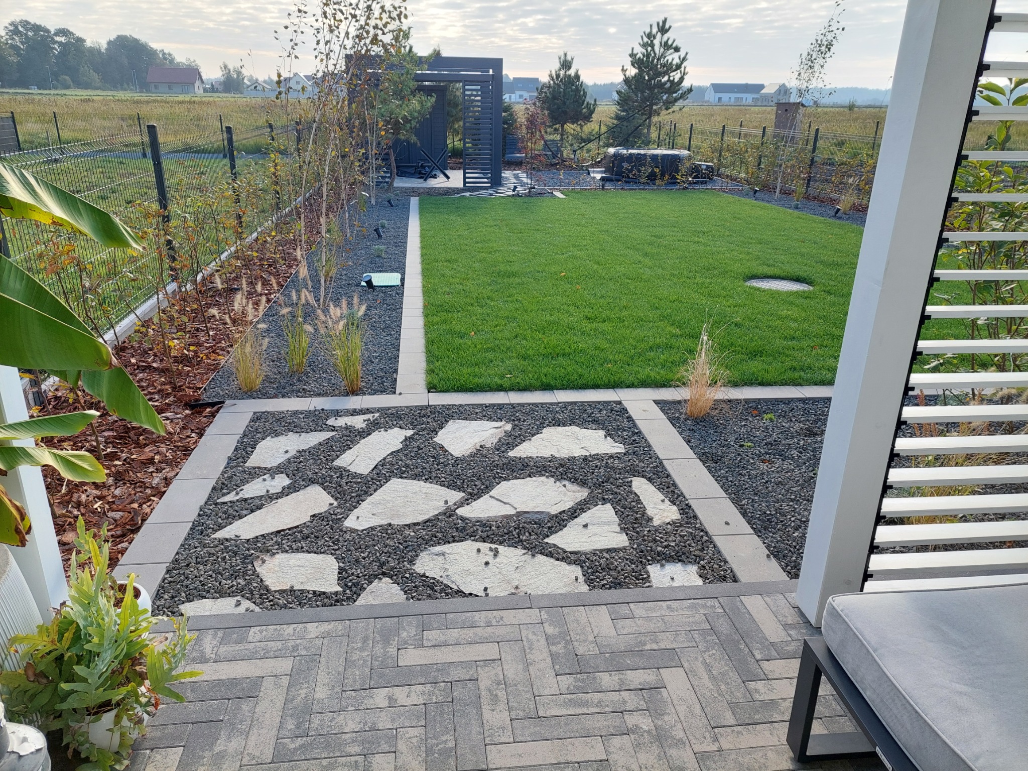 Nowoczesny ogród z równo przystrzyżonym trawnikiem, ścieżką z nieregularnych kamieni otoczonych grysem, pergolą w tle i roślinnością ozdobną wzdłuż ogrodzenia.