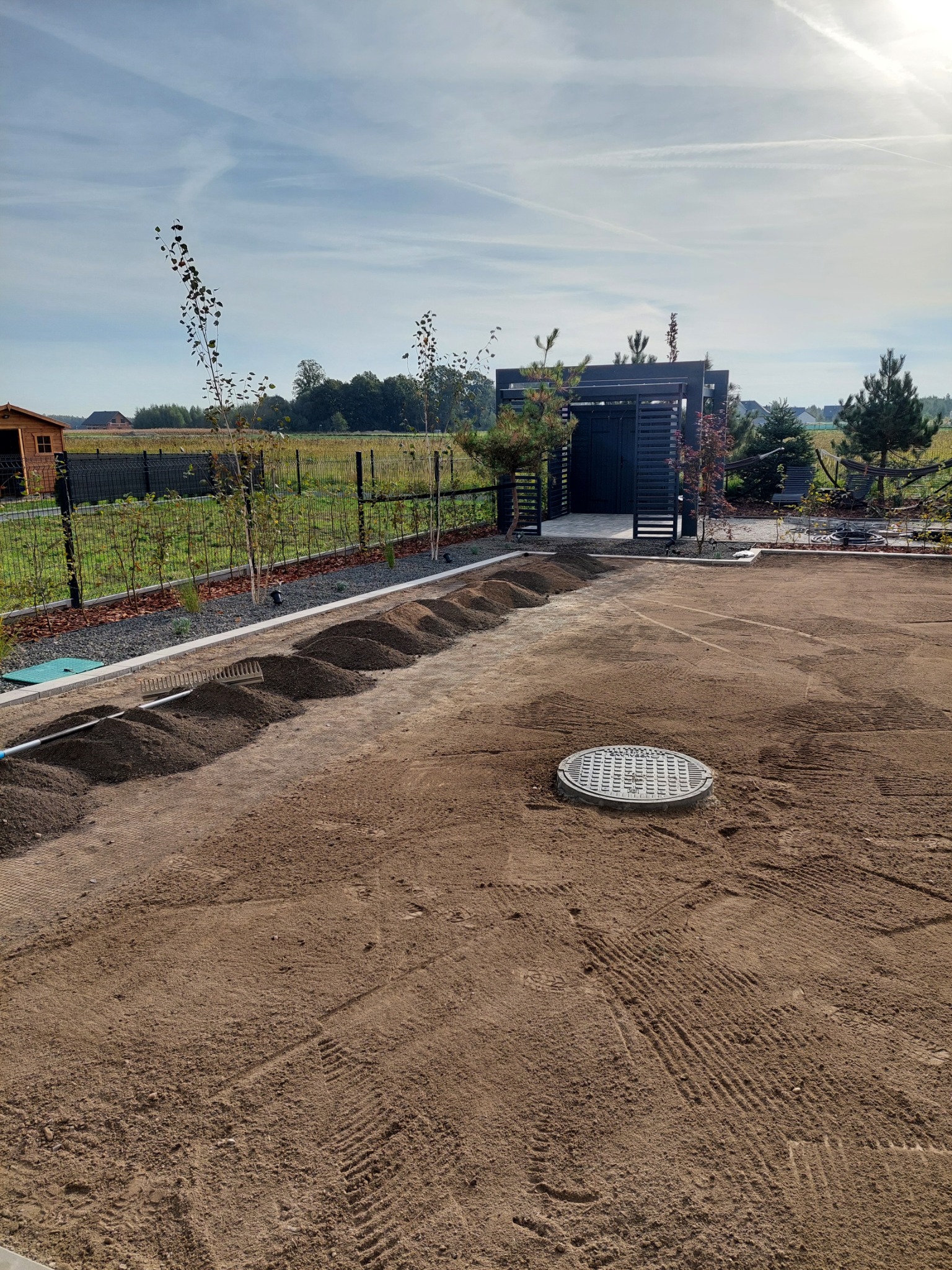 Przygotowanie podłoża pod trawnik z rolki: wyrównany teren z widocznymi śladami po maszynach, ułożone kopce ziemi wzdłuż rabaty z młodymi drzewkami, studzienka kanalizacyjna na pierwszym planie.