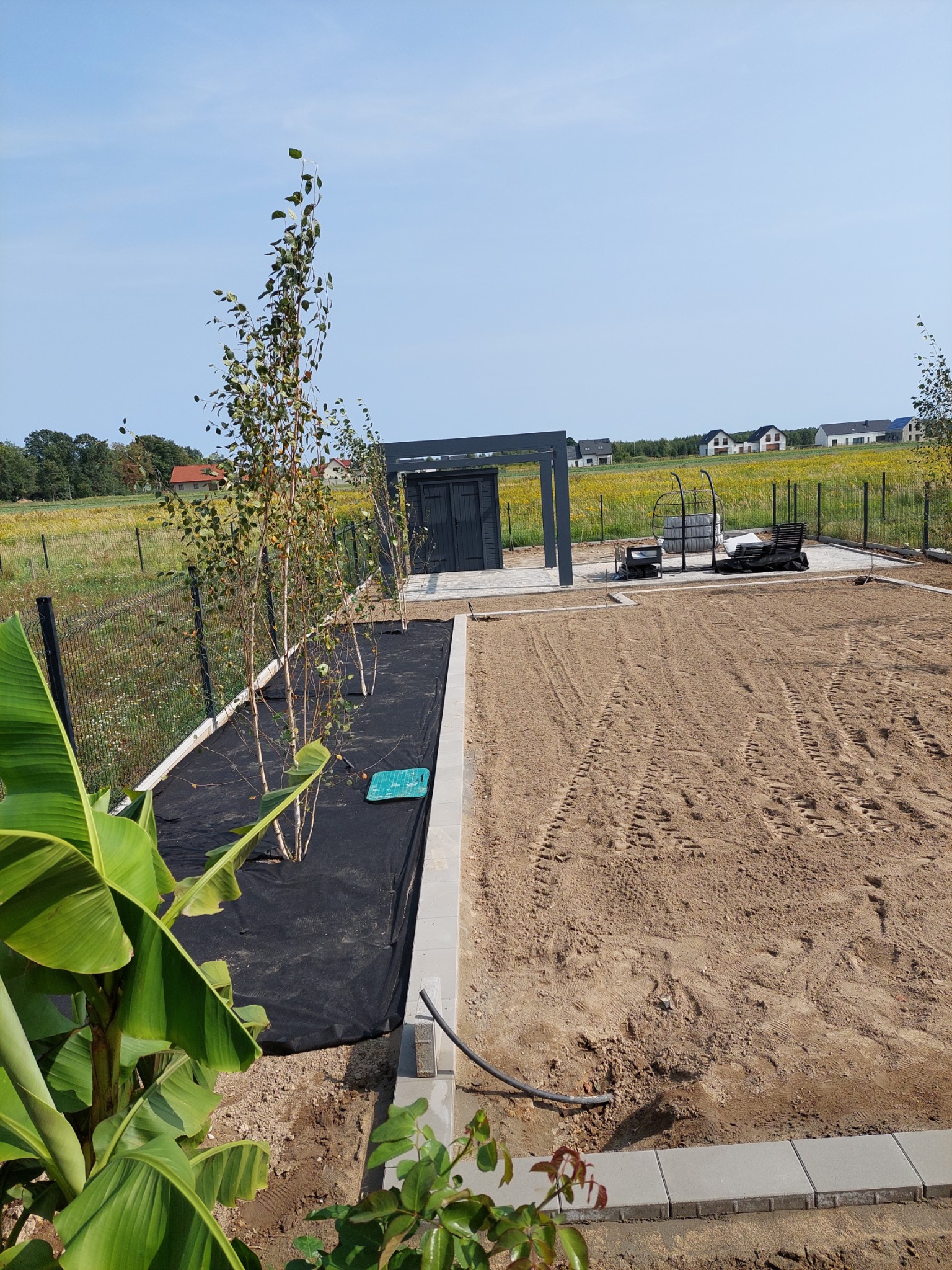 Przygotowany teren pod ogród: wyrównana ziemia z widocznymi śladami opon, obrzeża z kostki, posadzone młode drzewka na czarnej macie, pergola w tle.