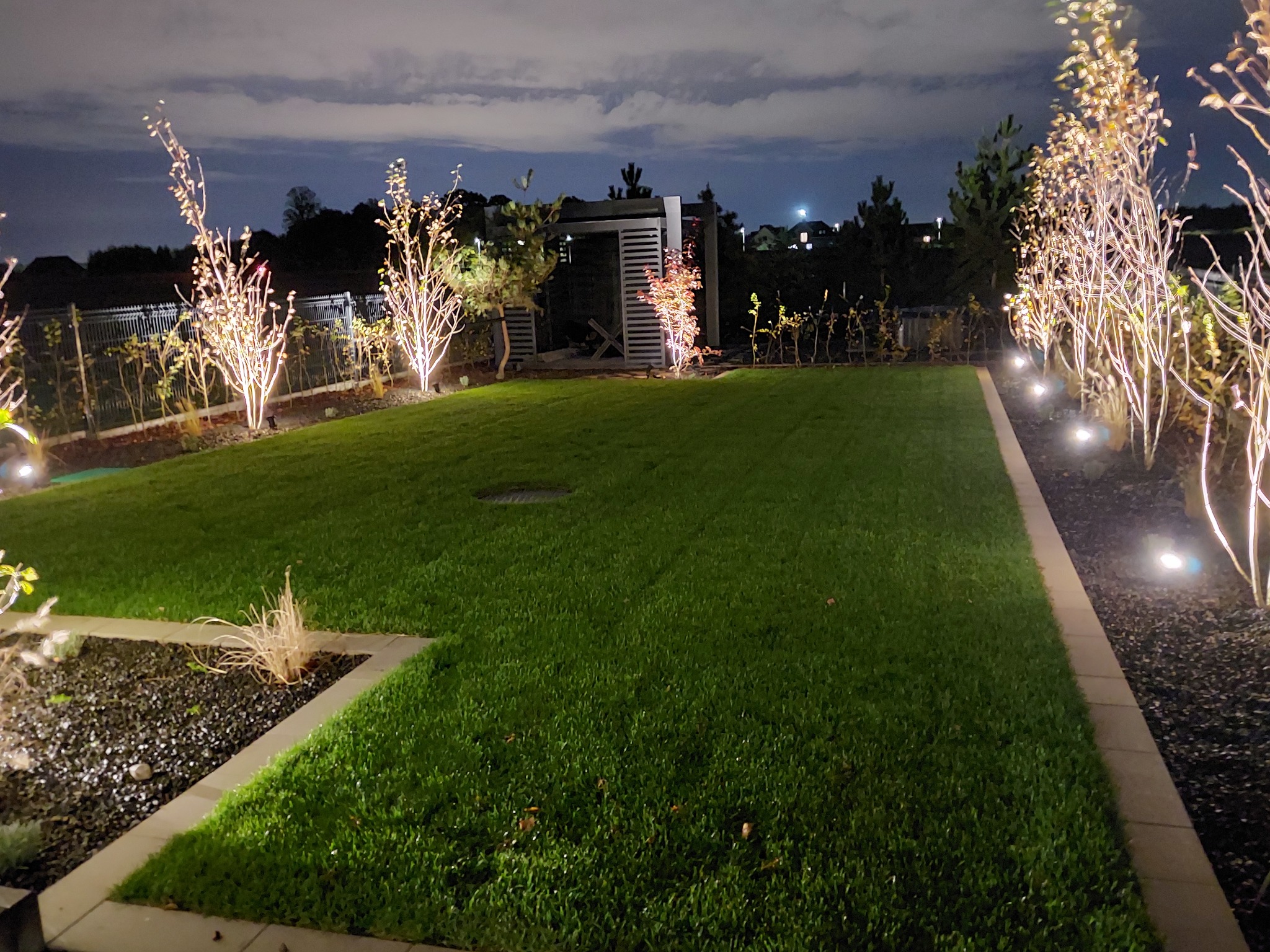 Ogród nocą z równo przystrzyżonym trawnikiem, podświetlonymi drzewami i pergolą w tle, oświetlenie punktowe podkreśla detale rabat z czarnym grysem.