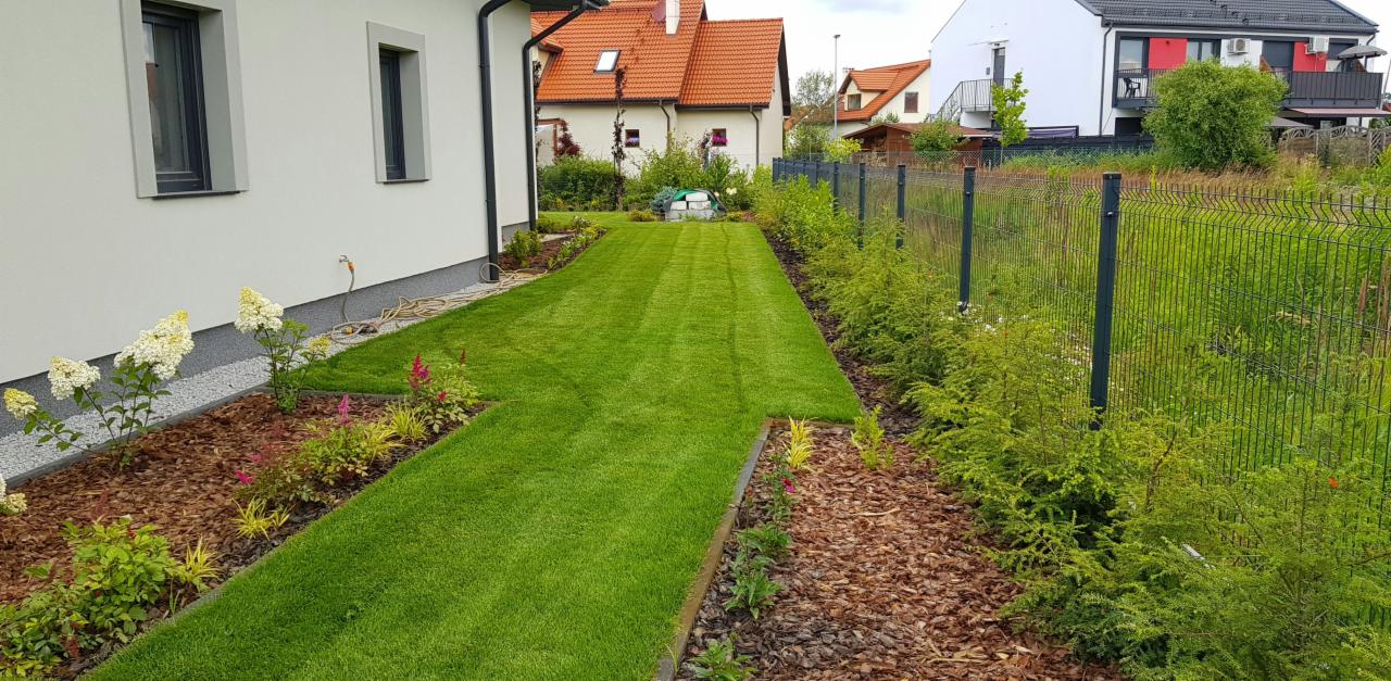 Zadbany trawnik przydomowy z równo przystrzyżoną trawą, obrzeżami z kory i nasadzeniami kwiatów wzdłuż domu i ogrodzenia panelowego.