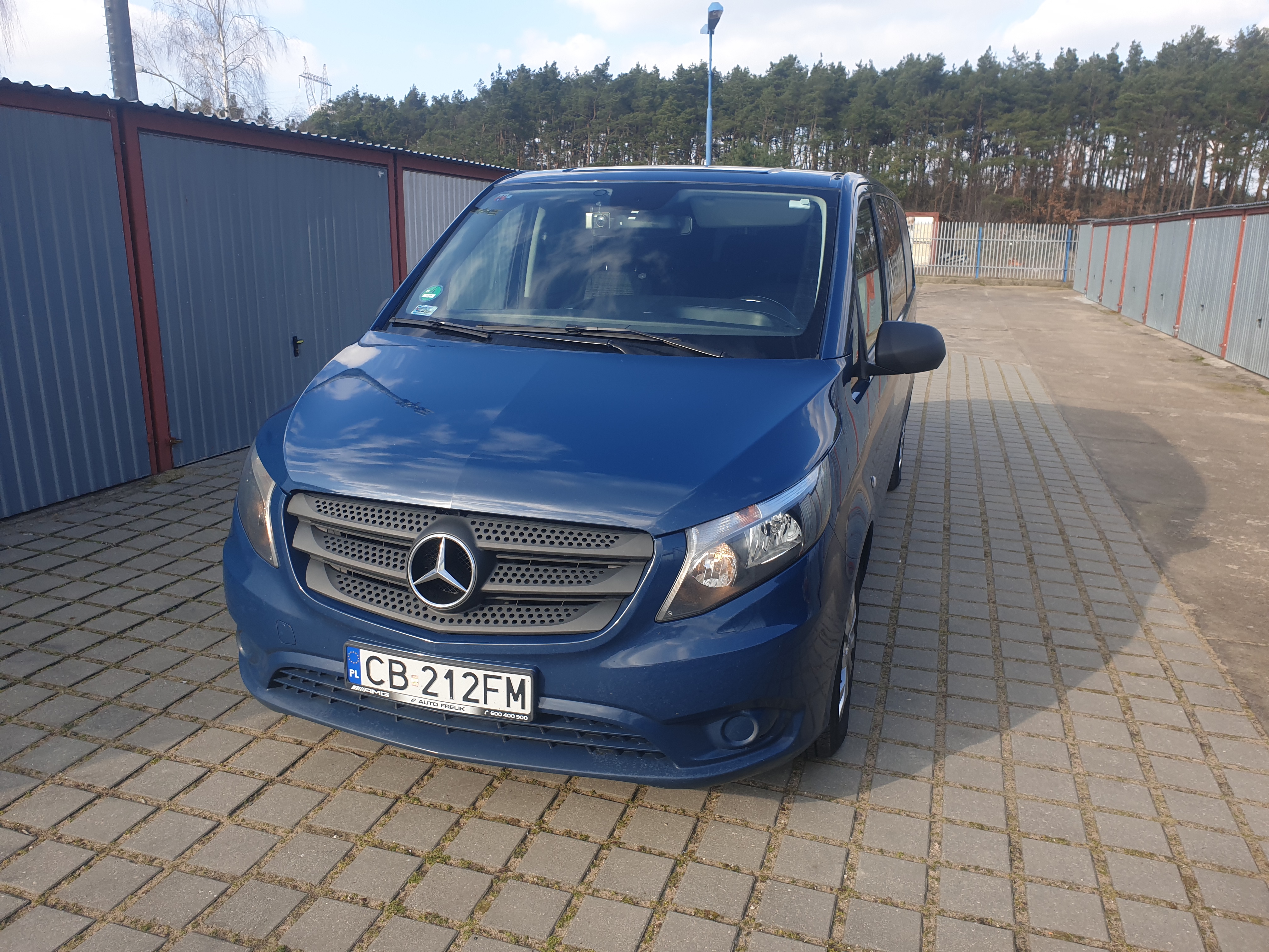 Niebieski Mercedes Vito zaparkowany na brukowanym placu, na tle rzędu garaży i drzew.