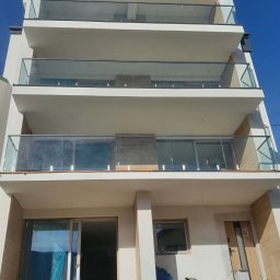 PEDROSTAL Mariusz Szyjka - Wielopiętrowy budynek z balkonami zabezpieczonymi szkłem i metalem, widok z dołu na błękitne niebo. Część balkonów jeszcze w trakcie montażu, widoczne materiały budowlane.