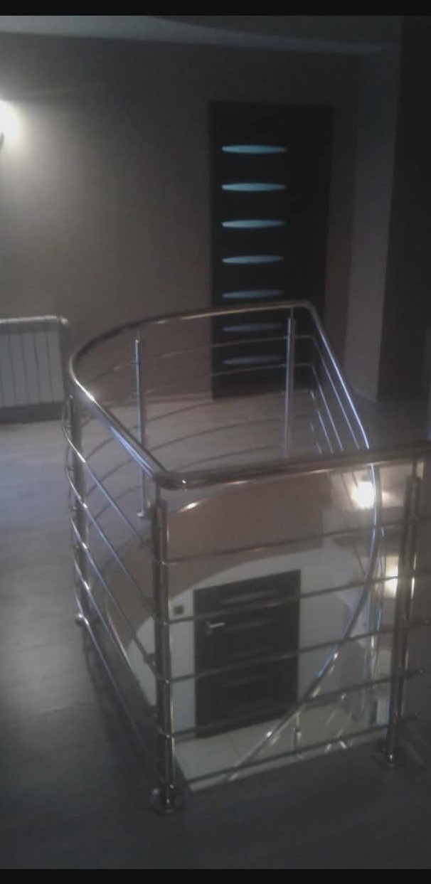 Spiralna, metalowa konstrukcja schodów z połyskującymi, cienkimi prętami, widoczna w słabo oświetlonym wnętrzu. W tle drzwi z poziomymi przeszkleniami.