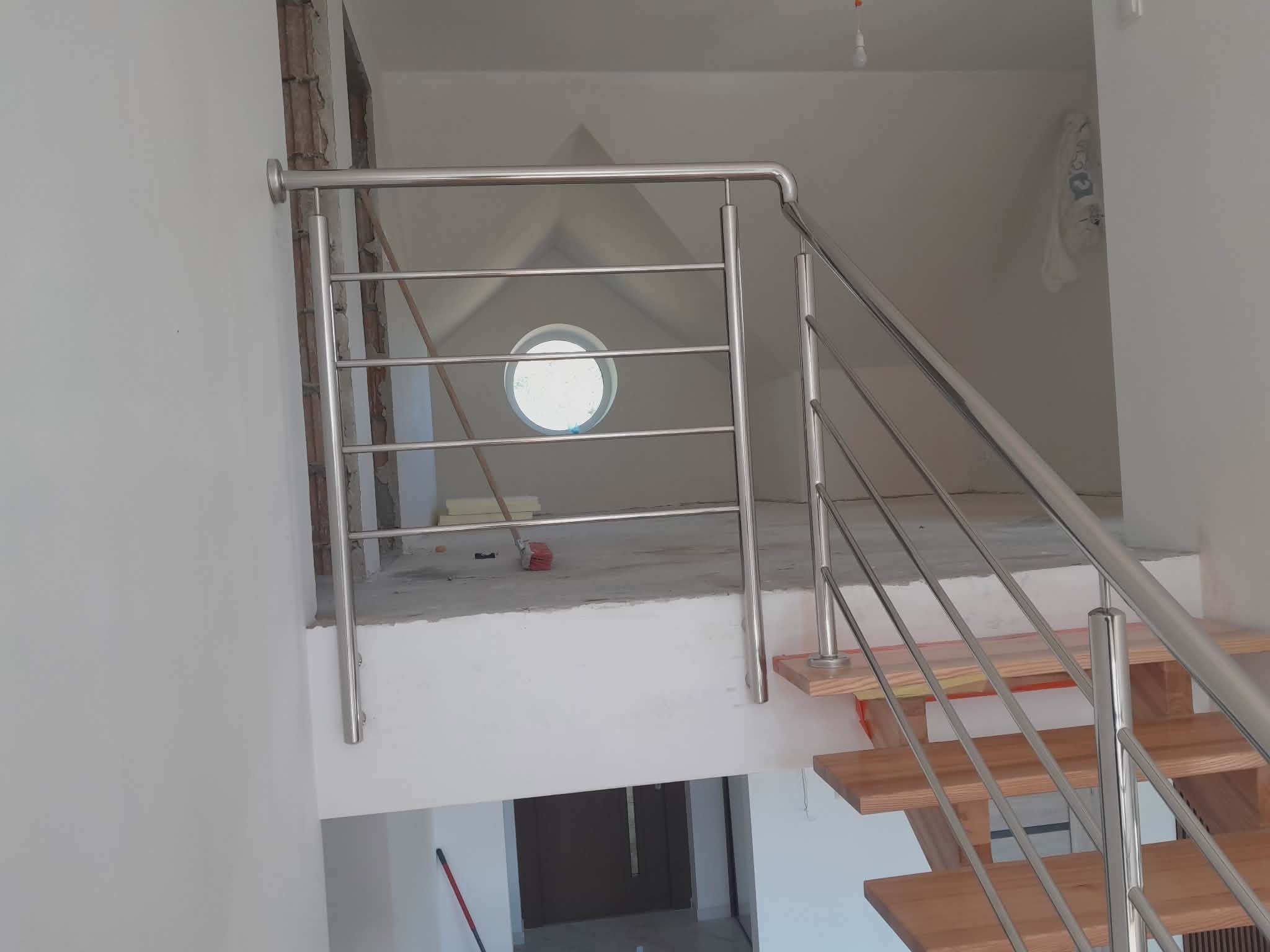 Nowoczesna, metalowa poręcz schodowa z poziomymi prętami, zamontowana w jasnym wnętrzu z drewnianymi schodami. Widoczne okrągłe okno w tle.