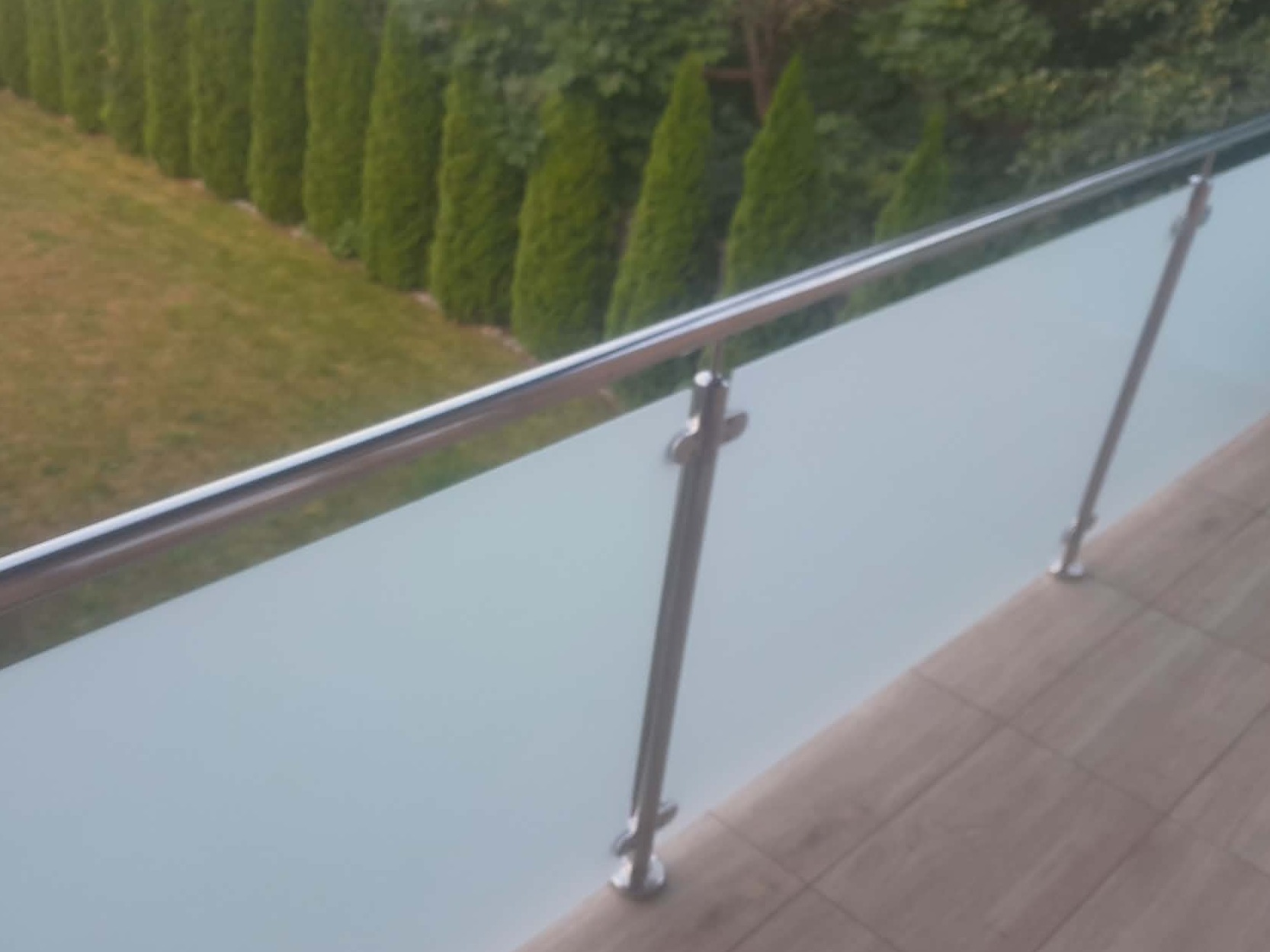 Szklana balustrada z elementami ze stali nierdzewnej na tarasie z widokiem na ogród z tujami. Nowoczesny design, jasne kolory, naturalne światło.