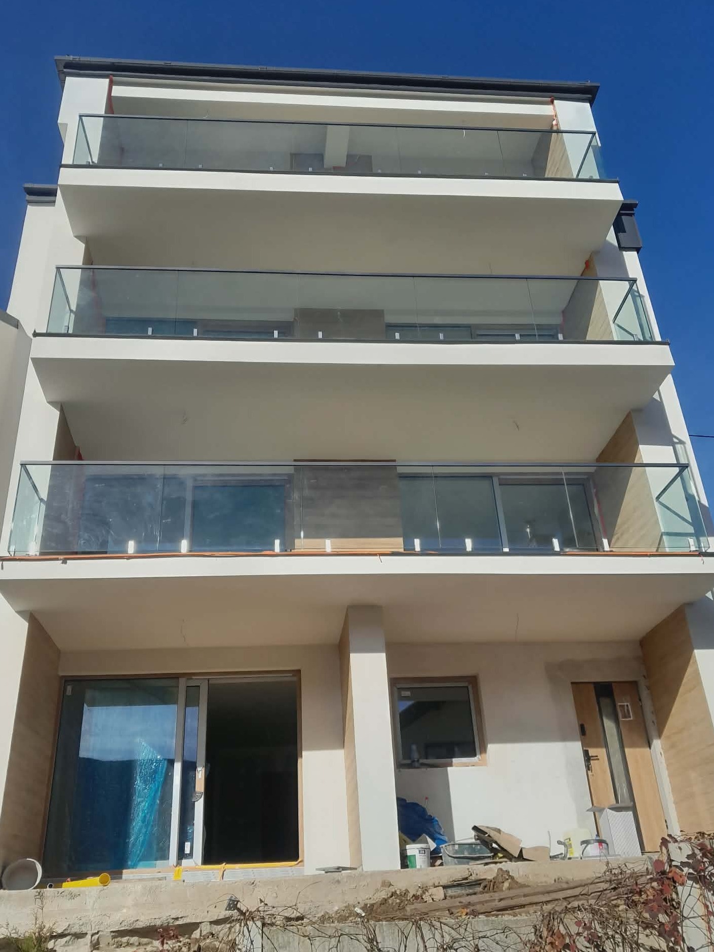 Wielopiętrowy budynek z balkonami zabezpieczonymi szkłem i metalem, widok z dołu na błękitne niebo. Część balkonów jeszcze w trakcie montażu, widoczne materiały budowlane.
