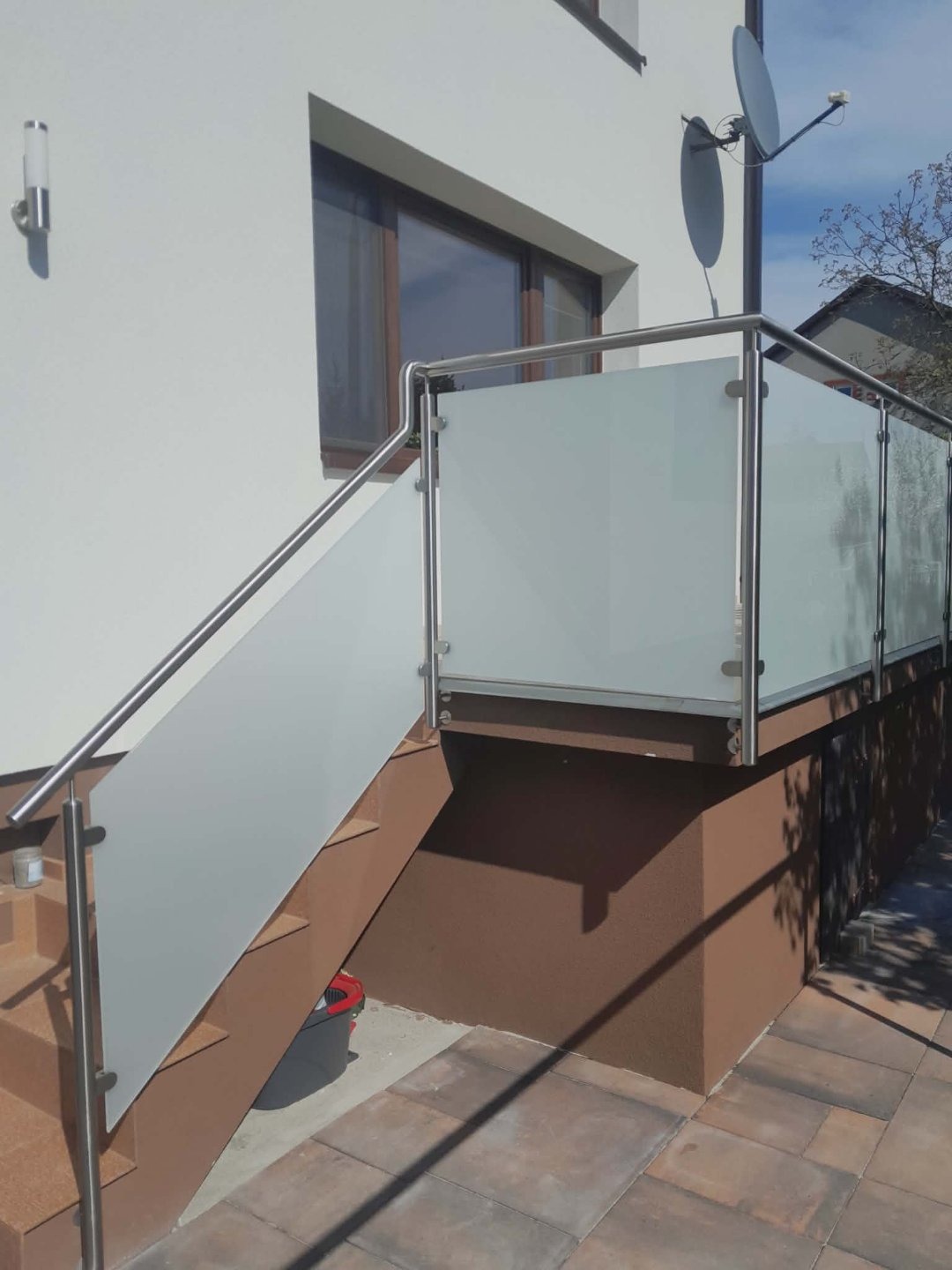 Zewnętrzna balustrada ze szkła matowego i stali nierdzewnej, zamontowana przy schodach wejściowych do domu. Widoczne detale mocowania i satelita na elewacji.