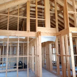 Surowa konstrukcja drewniana domu w trakcie budowy, widoczne ściany szkieletowe, więźba dachowa i czarne worki na śmieci w tle.