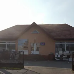 Budynek sklepu z materiałami budowlanymi, z ekspozycją płytek i paneli przed wejściem oraz zaparkowanym samochodem dostawczym i osobowym.