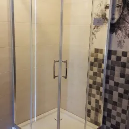 Nowoczesna kabina prysznicowa z chromowanymi profilami i przezroczystym szkłem, zamontowana w łazience z beżowymi płytkami i dekoracyjną mozaiką z motywem twarzy.