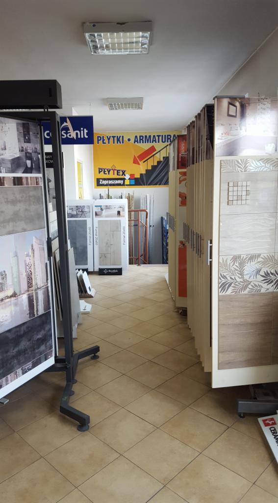 Wnętrze sklepu z płytkami ceramicznymi, ekspozytory z różnymi wzorami i kolorami płytek, w tle baner reklamowy 'Płytki Armatura'.