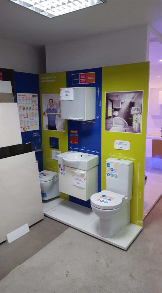 Ekspozycja w sklepie z armaturą łazienkową: biała toaleta z kolorowymi wzorami na desce, umywalka z szafką i wisząca szafka, wszystko na tle niebiesko-zielonej ściany z informacjami o produktach.