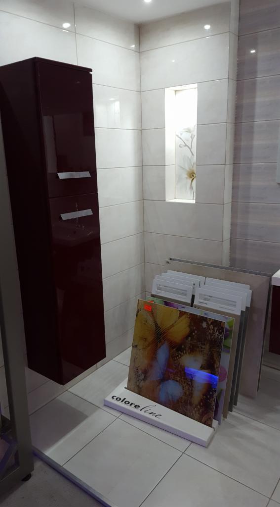 Ekspozycja płytek ceramicznych w salonie łazienek, widoczny wysoki, ciemny słupek łazienkowy z szufladami i wnęka z dekoracją na ścianie wyłożonej jasnymi płytkami.