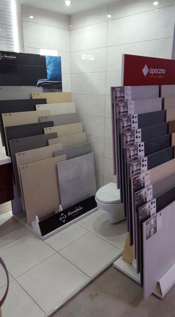 Ekspozycja płytek ceramicznych marek Nowa Gala i Opoczno w salonie, z widoczną toaletą w tle, prezentująca różne odcienie szarości, beżu i bieli.