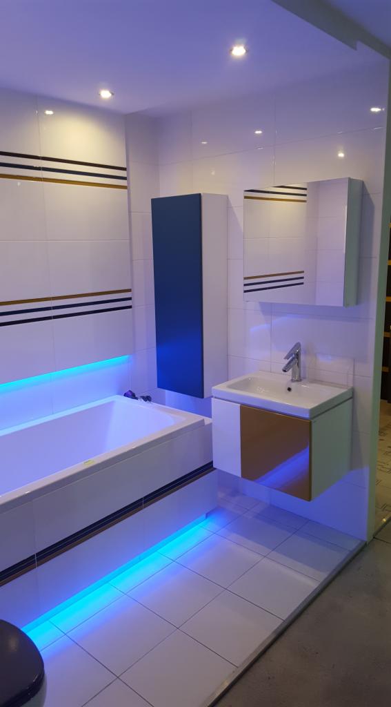 Nowoczesna łazienka z podświetleniem LED, białe płytki z dekoracyjnymi paskami, szafka umywalkowa z drewnianym akcentem i niebieska szafka wisząca.