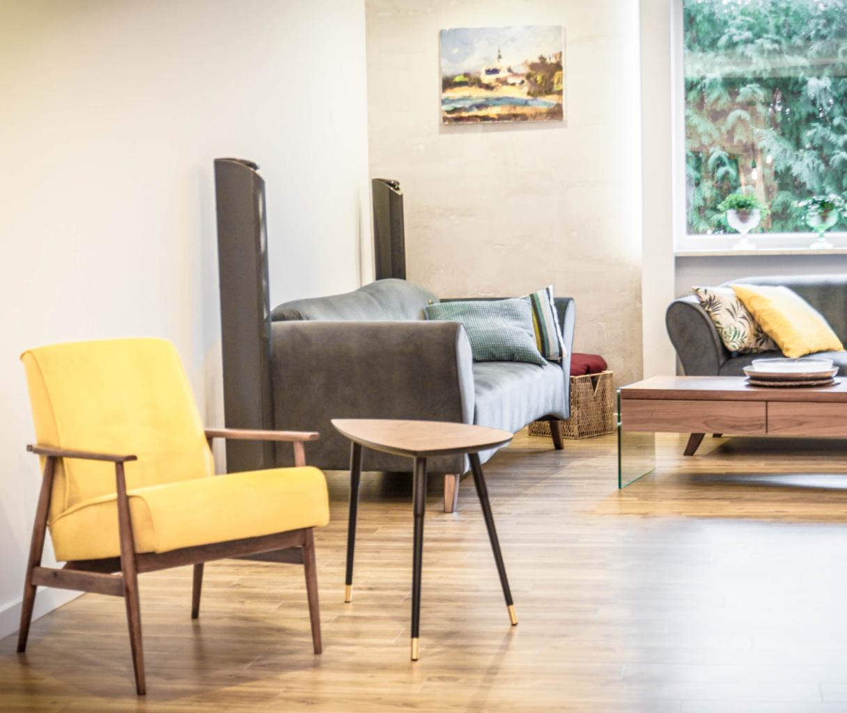 Salon z żółtym fotelem, szarymi sofami, drewnianym stolikiem kawowym z elementami szkła i obrazem nad sofą, oświetlony naturalnym światłem z okna z widokiem na zieleń.