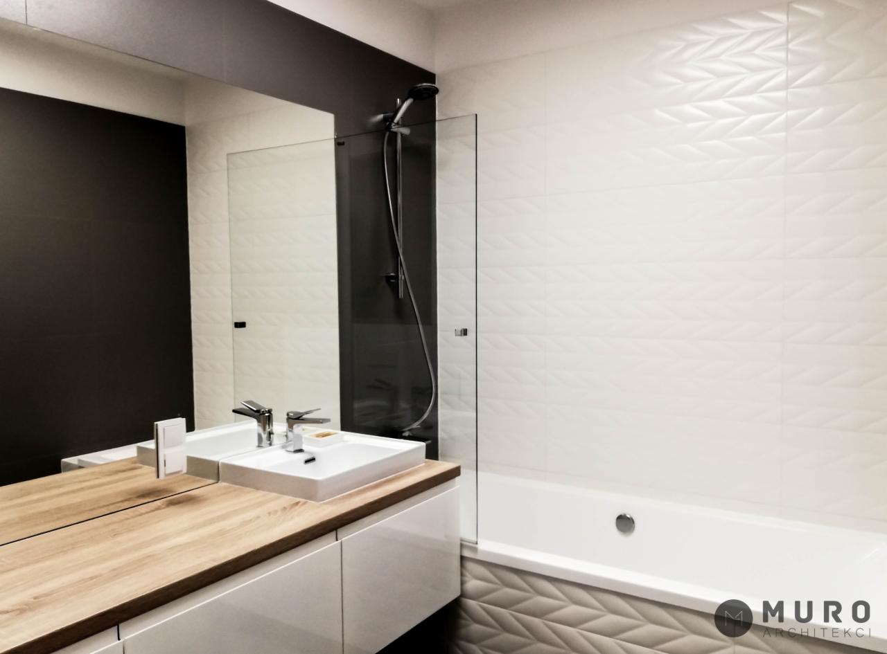 Nowoczesna łazienka z drewnianym blatem, białą umywalką i baterią, lustrem odbijającym ciemną ścianę oraz białą ścianą z teksturą 3D i wanną, widoczny fragment logo architektonicznego w prawym...