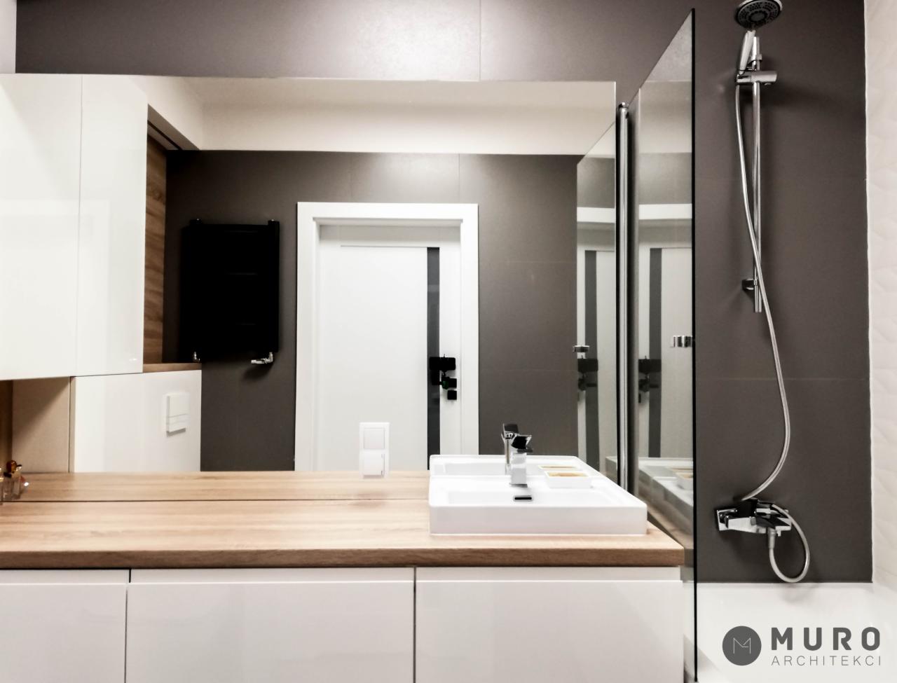 Nowoczesna łazienka z drewnianym blatem, białą zabudową meblową, umywalką nablatową i szarą ścianą z prysznicem typu walk-in, widoczne logo firmy architektonicznej w prawym dolnym rogu.