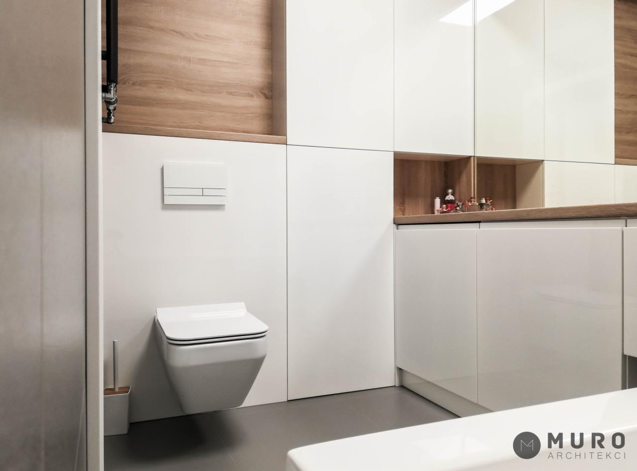 Minimalistyczna łazienka z podwieszaną toaletą, białą zabudową meblową z drewnianymi akcentami i dekoracyjnymi drobiazgami w otwartej półce.