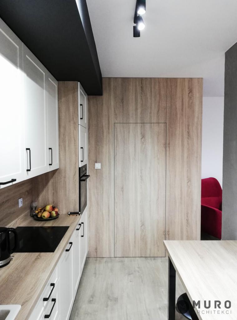 Nowoczesna kuchnia z białymi szafkami, drewnianym blatem i ukrytymi drzwiami, oświetlona punktowymi lampami, z widocznym fragmentem czerwonego fotela w tle.