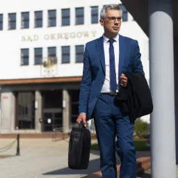 Mężczyzna w niebieskim garniturze i okularach, trzymający czarną torbę i marynarkę, stoi na zewnątrz budynku Sądu Okręgowego.