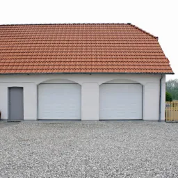 Biały budynek z dwoma segmentowymi bramami garażowymi z panelami, czerwony dach z dachówki, szare drzwi wejściowe, żwirowe podłoże.