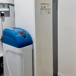 Biała pompa ciepła Atlantic stoi obok zmiękczacza wody z niebieską pokrywą w pomieszczeniu technicznym z białymi ścianami i panelami na podłodze.