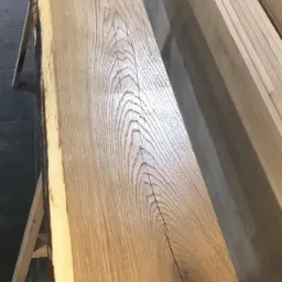 Surowa deska dębowa z korą na krawędzi, umieszczona na drewnianych kozłach, ukazująca wyraźny rysunek słojów i naturalne pęknięcia drewna.